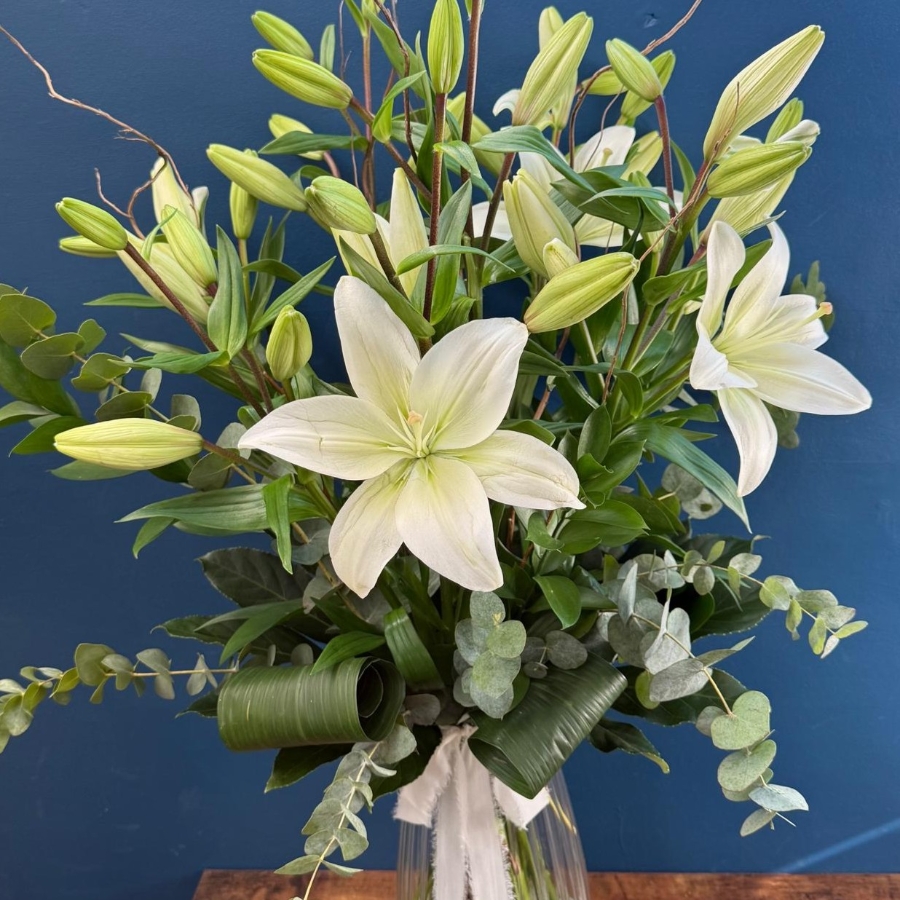 Tall white lily arrangement in glass vase with foliage and decorative twigs