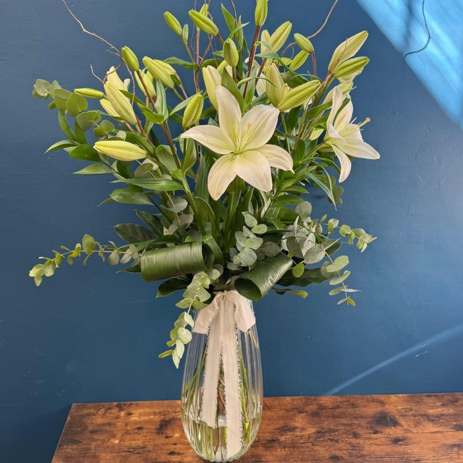Tall white lily arrangement in glass vase with foliage and decorative twigs